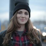 Young woman in a beanie smiling outdoors with urban backdrop, exuding warmth.